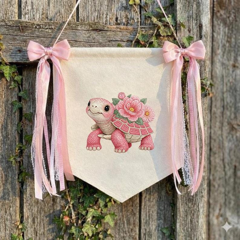 May include: A cream banner with a pink turtle illustration, decorated with pink flowers and bows. The banner is suspended by pink ribbons. The banner is hanging on a wooden surface. The overall design is charming and decorative.