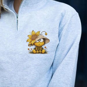 May include: Light gray sweatshirt with a quarter-zip collar. The sweatshirt features a whimsical cartoon bee wearing a sunflower hat. The bee is sitting in a patch of grass with small orange flowers. The bee has a surprised expression.