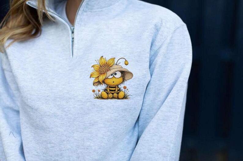 May include: Light gray sweatshirt with a quarter-zip collar. The sweatshirt features a whimsical cartoon bee wearing a sunflower hat. The bee is sitting in a patch of grass with small orange flowers. The bee has a surprised expression.