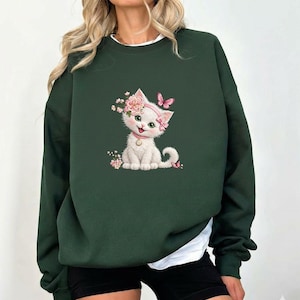 May include: Dark green sweatshirt featuring a white kitten illustration adorned with pink flowers and a butterfly. The kitten wears a pink collar and a floral headband. The sweatshirt has a crew neck and long sleeves, with a white undershirt peeking out.