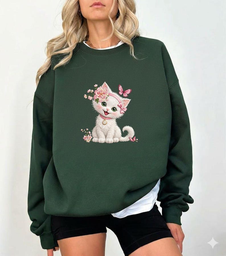 May include: Dark green sweatshirt featuring a white kitten illustration adorned with pink flowers and a butterfly. The kitten wears a pink collar and a floral headband. The sweatshirt has a crew neck and long sleeves, with a white undershirt peeking out.