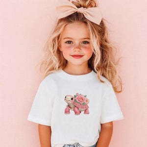 May include: A white t-shirt with a pink turtle design and flowers. The turtle has a pink shell and legs, and is decorated with pink flowers. The shirt is worn by a young child with blonde hair and a pink bow.