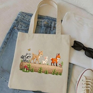May include: Beige tote bag featuring a woodland scene with a raccoon, fawn, rabbit, and fox on a log. The design includes green foliage, mushrooms, and a butterfly. The bag has two handles and is placed on a pair of blue jeans and a white t-shirt.