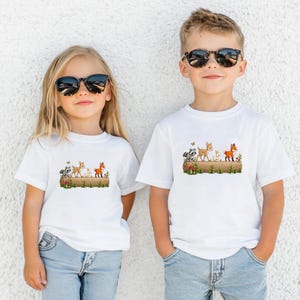 May include: Two children wearing white t-shirts with a woodland animal design. The shirts feature a raccoon, deer, rabbit, and fox on a log with mushrooms and grass. Both children are wearing sunglasses and blue jeans.
