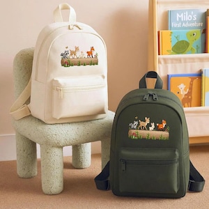 May include: Two backpacks, one cream-colored and one dark green, are displayed. Each backpack features a front pocket and a top handle. The cream backpack is on a light green chair. Both backpacks have an embroidered woodland animal design.