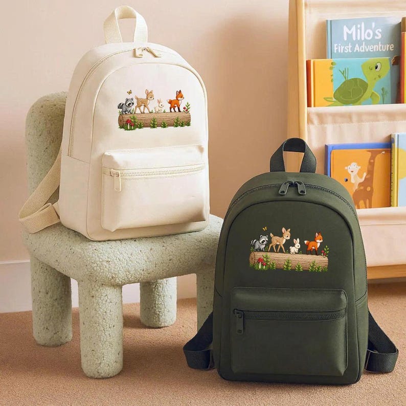 May include: Two backpacks, one cream-colored and one dark green, are displayed. Each backpack features a front pocket and a top handle. The cream backpack is on a light green chair. Both backpacks have an embroidered woodland animal design.