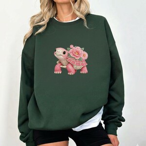 May include: Dark green sweatshirt with a pink turtle and floral design. The turtle has a smiling face and is adorned with pink flowers. The sweatshirt has a crew neck and long sleeves.