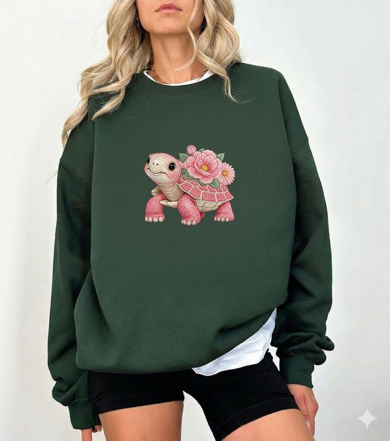 May include: Dark green sweatshirt with a pink turtle and floral design. The turtle has a smiling face and is adorned with pink flowers. The sweatshirt has a crew neck and long sleeves.