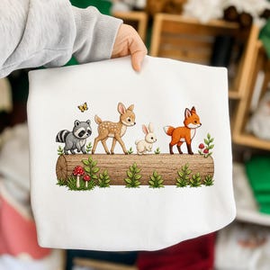 May include: White sweatshirt featuring a woodland scene with a raccoon, deer, rabbit, and fox on a log. The design includes a yellow butterfly, red mushrooms, and green foliage. The illustration has a cartoon style.
