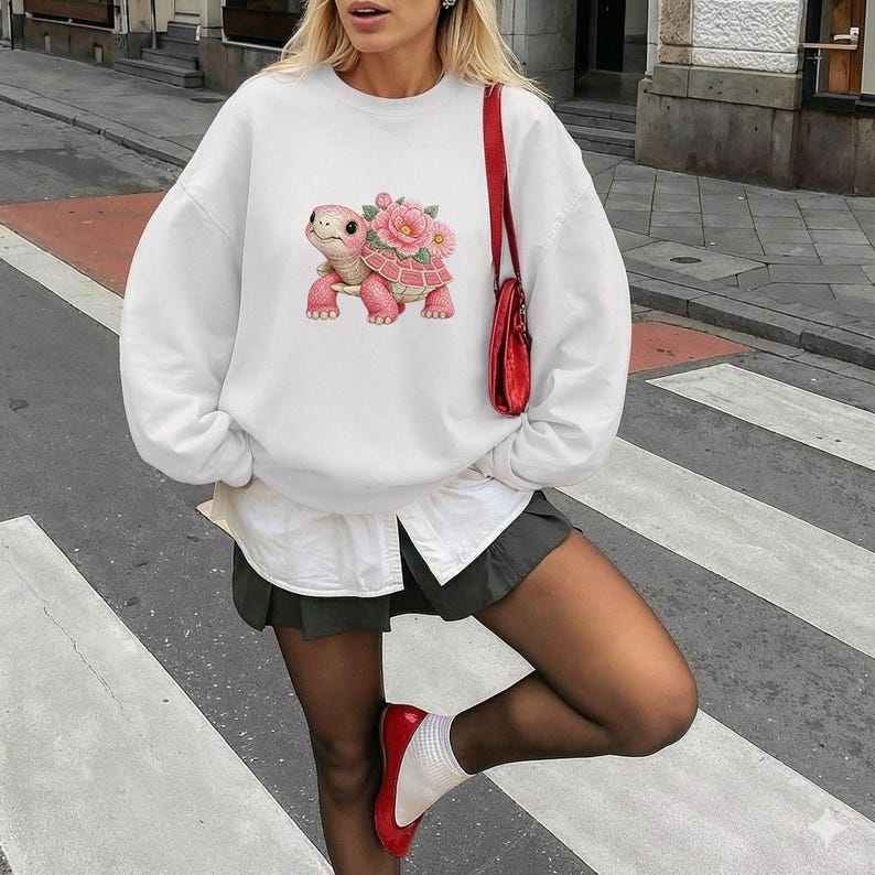 May include: White sweatshirt with a pink turtle and floral design. The turtle has a pink shell and is surrounded by pink flowers. The sweatshirt is a casual style.