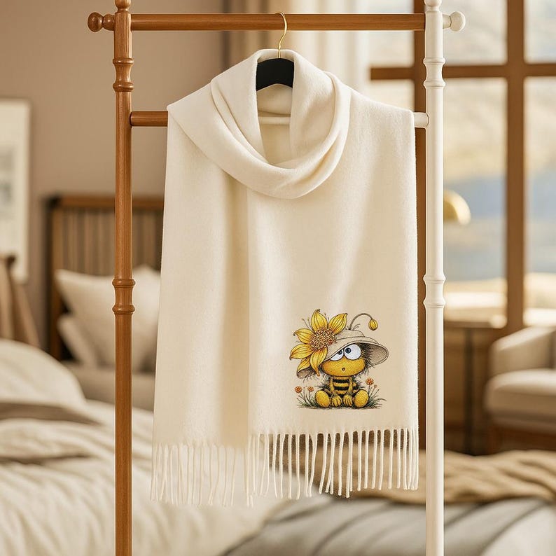 May include: Cream-colored scarf with a fringed edge, hanging on a wooden rack. The scarf features a whimsical illustration of a bee wearing a hat and a sunflower. The scarf is made of a soft material.