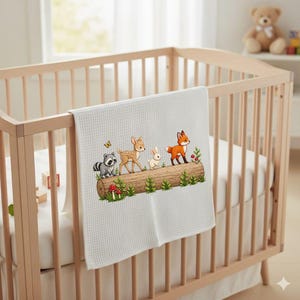 May include: A white baby blanket with a woodland animal design. The blanket features a raccoon, deer, rabbit, and fox standing on a log with mushrooms and greenery. The blanket is displayed on a wooden crib.