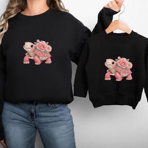 May include: Two black sweatshirts, one adult and one child size, each with a pink turtle design and floral details. The turtle has a pink shell and legs, and a flower crown. The sweatshirts are a matching set.