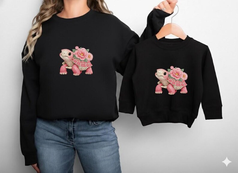 May include: Two black sweatshirts, one adult and one child size, each with a pink turtle design and floral details. The turtle has a pink shell and legs, and a flower crown. The sweatshirts are a matching set.