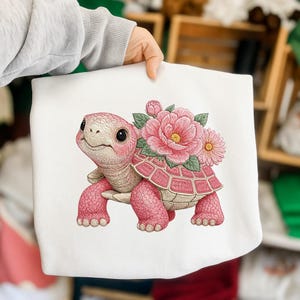 May include: White sweatshirt with a pink turtle and floral design. The turtle has a pink shell, legs, and head, with a bouquet of pink flowers on its back. The turtle has a smiling expression.