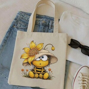 May include: Beige tote bag featuring a cartoon bee wearing a sunflower hat. The bee is yellow and black, sitting in a field of small orange flowers. The bag has two beige handles and is placed on top of blue jeans and a white t-shirt.