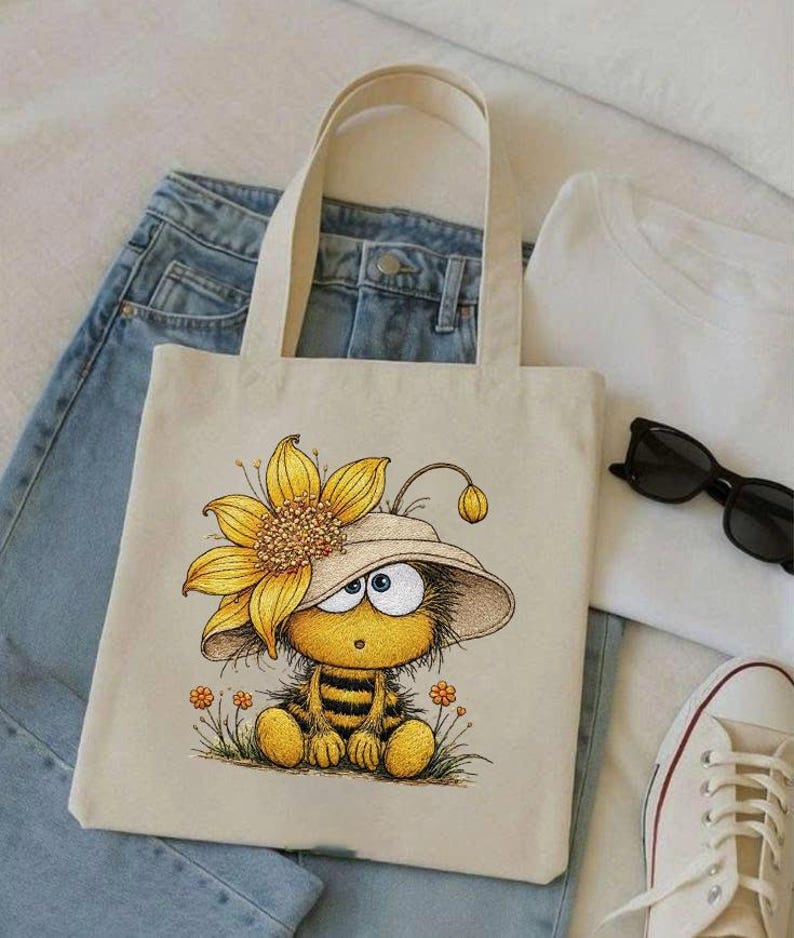 May include: Beige tote bag featuring a cartoon bee wearing a sunflower hat. The bee is yellow and black, sitting in a field of small orange flowers. The bag has two beige handles and is placed on top of blue jeans and a white t-shirt.