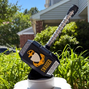 May include: A black hammer with a silver handle and a silver-toned decorative pattern on the sides. The hammer features a gold-colored anchor and shield emblem, with the word SWAGGER printed in yellow capital letters on the side of the head.