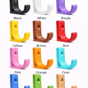 Peut inclure: Onze crochets muraux de différentes couleurs : noir, blanc, violet, jaune, marron, rouge, rose, orange, cyan, bleu, vert et chêne clair. Chaque crochet a une forme en L avec un petit trou. Le texte "Les couleurs peuvent varier légèrement en raison de l'impression" est en bas.