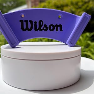 May include: A purple Wilson tennis visor sits atop a white, round container. The visor has the Wilson logo in black. The container is on a white surface, with green foliage in the background. The visor is designed to shield the eyes from the sun.