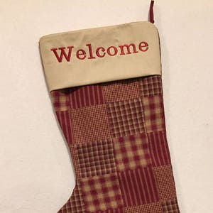May include: A Christmas stocking with a patchwork design in red and tan. The top cuff is tan with the word "Welcome" embroidered in red. The stocking has a loop for hanging.