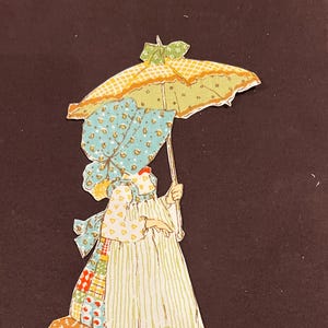 Op de afbeelding: Een vintage papieren poppenknipsel van een meisje in een patchworkjurk en een blauwe muts, die een geel-groene gestreepte paraplu vasthoudt.