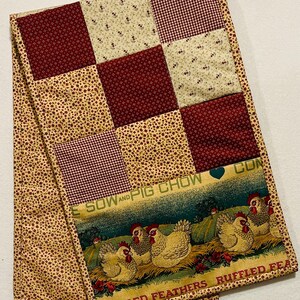 Può includere: Runner da tavolo trapuntato con un design patchwork di tessuti bordeaux e color crema. Il bordo inferiore presenta una scena pastorale con polli e il testo "E SOW AND PIG CHOW" e "RUFFLED FEATHERS".