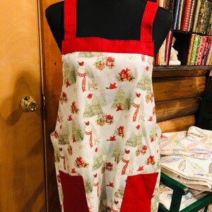 Puede incluir: Un delantal blanco con ribete rojo y bolsillos rojos. El delantal tiene un patrón invernal con muñecos de nieve, cardenales y regalos.