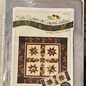 Op de afbeelding: Een quiltpatroon voor een Woodland Stars quilt met een bruin, groen en rood kleurenschema. Het patroon is ontworpen door Dodi Lee Poulsen en maakt deel uit van de Two Sisters at Squirrel Hollow collectie. Het patroon bevat instructies voor een tafelloper en een charm pack-vriendelijke quilt.