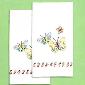 May include: Two white decorative towels with embroidered butterfly designs. The butterflies feature yellow, pink, and blue wings. A row of small flowers in red, pink, and green adorns the bottom of each towel. The towels are displayed on a green background.
