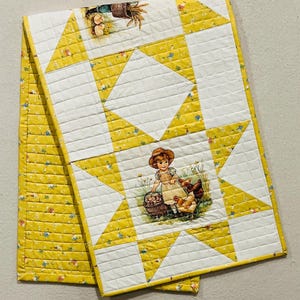 May include: A quilted fabric runner featuring a yellow and white star pattern. The design includes a central illustration of a child in a straw hat holding a basket near chickens, with additional small floral and farm-themed motifs throughout.