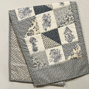 Puede incluir: Un edredón hecho a mano con un diseño de patchwork. Presenta cuadrados de color crema bordados con motivos de casitas para pájaros. El edredón tiene un borde y un reverso con estampado floral azul marino. La combinación de colores es crema y azul marino.