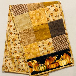 May include: A patchwork table runner with a fall theme. The runner is made of various fabrics in shades of brown, beige, and yellow. The runner features a border of pumpkins and leaves.