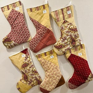 May include: Six quilted Christmas stockings numbered one through six, featuring patchwork designs in cream, red, and brown. Each stocking has a striped cuff and various floral or paisley patterns, with white hanging loops at the top.