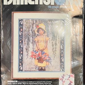 May include: A Dimensions No Count Cross Stitch kit titled Rebecca, featuring a portrait of a child in a yellow dress and bonnet holding a flower basket. The packaging includes text, a 14x16 inch frame size, and a 18x20 inch frame size with mat.