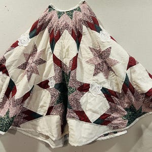 May include: A circular quilted tree skirt featuring a patchwork star pattern in cream, deep red, and dark green floral fabrics. The design includes white lace flower appliqués stitched onto the surface, with a central opening at the top.