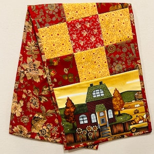May include: A quilted table runner with a patchwork design featuring red and yellow floral patterns. The runner has a border of red floral fabric and a center panel with a farmhouse scene featuring a house, trees, and pumpkins.