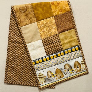 May include: A brown and yellow patchwork table runner with a beehive and honey jar design. The runner has a checkered pattern on the back.