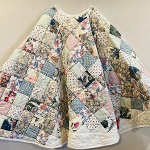 May include: A circular quilted tree skirt featuring a patchwork design of various floral and polka dot fabric squares in shades of pink, blue, green, and white. The skirt has a central opening and a solid white quilted border along the edge.
