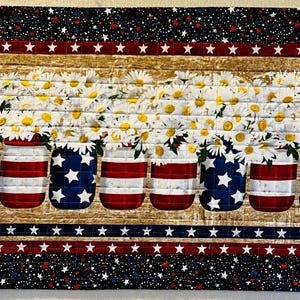 May include: A patriotic table runner featuring a design of mason jars with American flag patterns, filled with daisies. The runner has a black background with white stars, red and white stripes, and red corner accents.