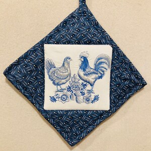 以下が含まれることがあります： 青と白の刺繍が施された鍋つかみで、雄鶏と雌鶏のデザインが施されています。鍋つかみは青と白のパターンで生地で作られています。