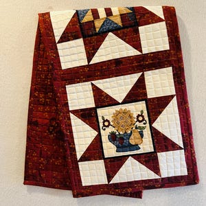 May include: Quilted table runner with a burgundy floral pattern and cream-colored star and square designs. The center features a floral arrangement in a blue vase with pears. The runner is approximately 40 inches long.
