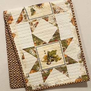 May include: A rectangular quilted table runner featuring a central star pattern with autumn-themed fabric prints of pumpkins, acorns, and leaves in shades of orange, green, and cream. The border has a brown and cream checkered pattern.