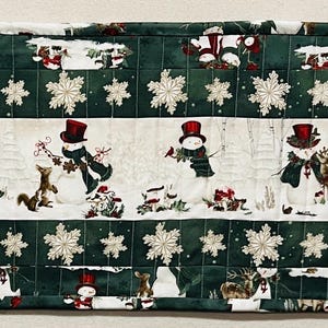 May include: A quilted Christmas table runner with a festive winter scene. The runner features a repeating pattern of snowmen, reindeer, and snowflakes on a white background, framed by a green border. The runner is approximately 48 inches long.