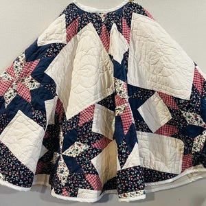 May include: A circular quilted tree skirt featuring a patchwork star pattern in navy blue, cream, and red plaid fabrics. The design incorporates floral print accents and solid cream panels with intricate stitched detailing throughout the fabric.