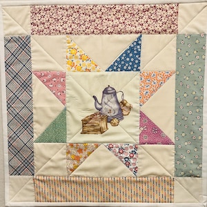 May include: A square fabric quilt block featuring a central cream panel with a printed illustration of a blue coffee pot, a wooden coffee grinder, and small pastries. The block is surrounded by a star pattern made of various floral and plaid fabrics.