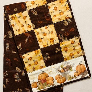 May include: A quilted table runner featuring a patchwork design with alternating squares of brown and cream fabric. The cream squares are decorated with orange pumpkins and fall leaves. The border has a repeating pattern of pumpkins, a birdhouse, and a bucket of harvest items.