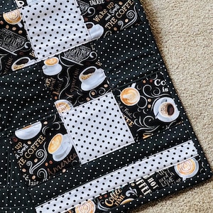 Puede incluir: Un mantel individual acolchado con temática de café con un diseño de patchwork. El mantel individual incorpora tela de lunares blancos y negros, junto con tela estampada con tazas de café, granos de café y frases como "Wake up and Smell the Coffee".