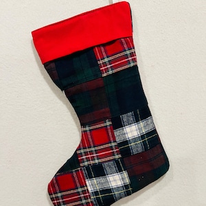May include: A Christmas stocking featuring a patchwork design of various plaid fabrics in shades of red, green, navy blue, and white. The top cuff is a solid bright red fabric, and a small white fabric loop is attached for hanging the item.