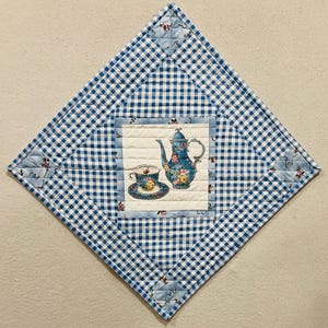 May include: A diamond-shaped quilted wall hanging featuring a blue and white gingham border. The center square displays a printed graphic of a blue floral teapot and a matching teacup on a saucer against a cream-colored background.